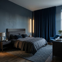 Une photographie d'une chambre à coucher moderne et moody aux murs bleu foncé. Le lit est habillé de draps en lin gris et en velours bleu, flanqué d'une table de chevet avec une lampe à gauche.