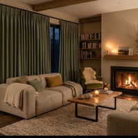 Intérieur d'un salon moderne et chaleureux à Bueil, Normandie, pendant l'hiver. De grands rideaux épais vert sauge ferment les fenêtres. La lumière douce de la cheminée et des lampes d'ambiance éclaire un canapé confortable avec un plaid